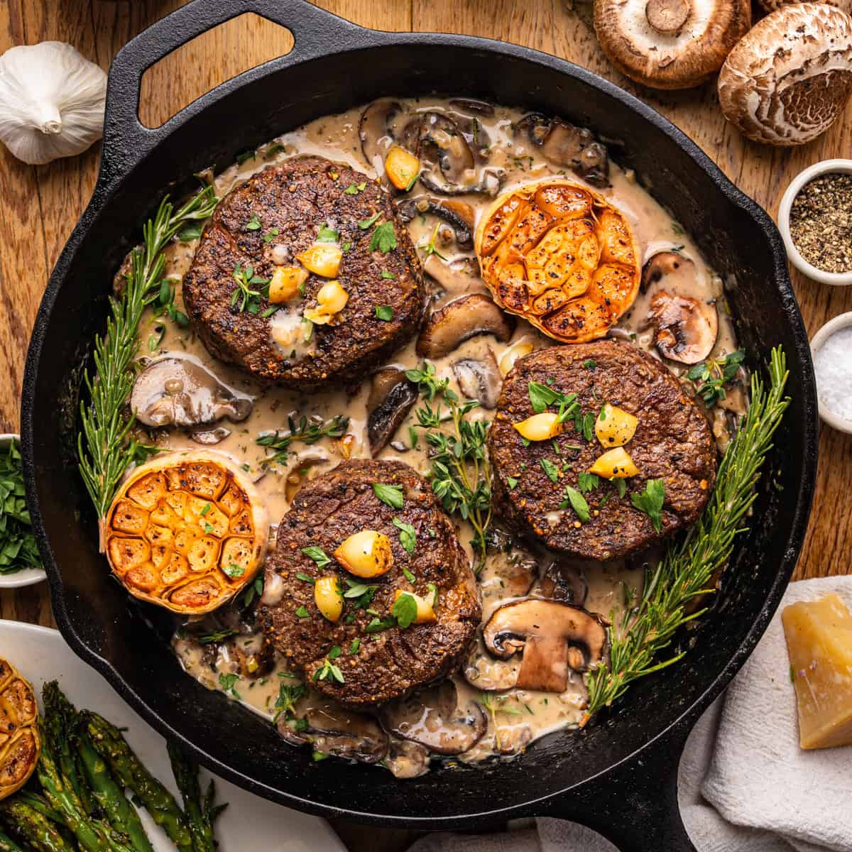 A cast iron skillet filled with three seared beef filet mignon steaks in creamy mushroom sauce, garnished with roasted garlic, fresh rosemary, and parsley, surrounded by mushrooms, garlic, salt, pepper, and a side of asparagus.