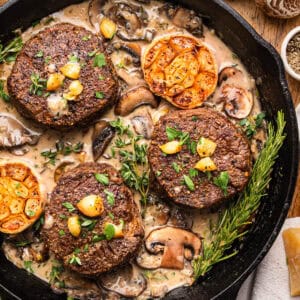 Three seared filet mignon steaks in a creamy mushroom sauce, garnished with parsley and roasted garlic cloves, in a black cast iron skillet with fresh rosemary and thyme.