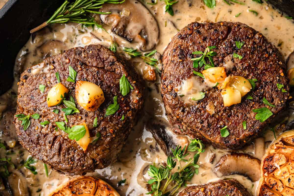 Two seared filet mignon steaks topped with garlic and fresh herbs sit in a creamy mushroom sauce, garnished with rosemary and parsley. Slices of roasted garlic and mushrooms are visible around the steaks.