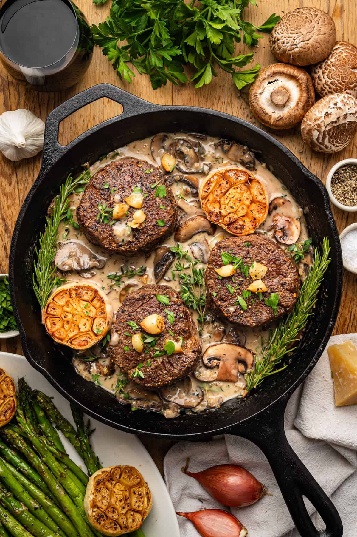 A cast iron skillet with three beef filet mignon medallions in creamy mushroom sauce, garnished with roasted garlic, herbs, and rosemary. Surrounding the skillet are fresh mushrooms, parsley, garlic, shallots, black pepper, and asparagus.