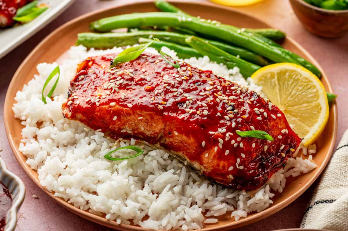A plate with white rice topped with gochujang salmon, glazed and sprinkled with sesame seeds and sliced green onions, served with green beans and a lemon slice.