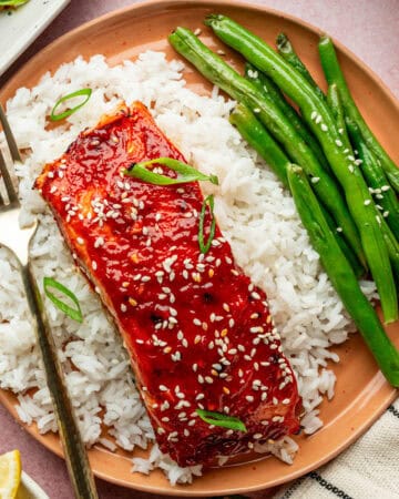 A plate with white rice topped with gochujang salmon glazed and sprinkled with sesame seeds and green onions, served with green beans. A fork rests on the side of the salmon.