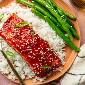 A plate with white rice, green beans, and gochujang salmon fillet glazed and topped with sesame seeds and sliced green onions. A fork rests on the plate next to a striped napkin.