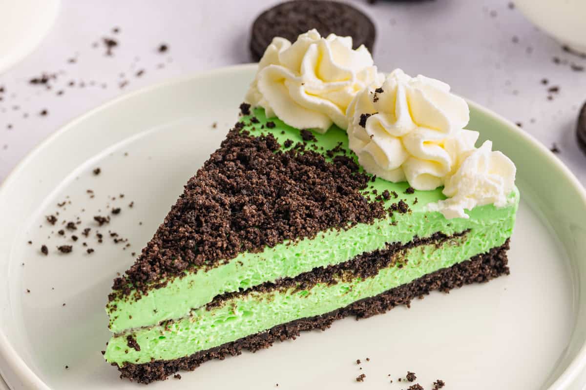 Grasshopper Cheesecake A slice of green mint Grasshopper cheesecake with a chocolate cookie crust and crumbs, topped with whipped cream, on a white plate.