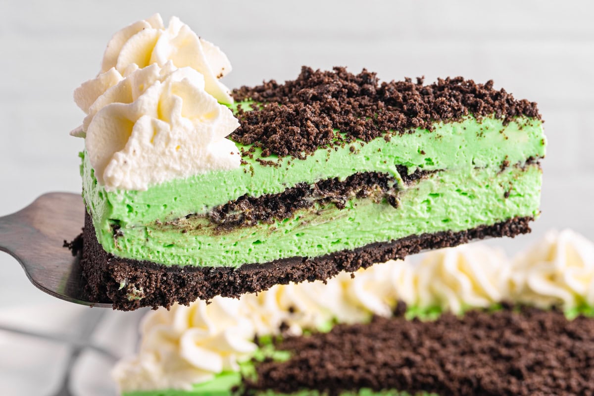 Grasshopper Cheesecake A close-up of a slice of Grasshopper Cheesecake with a chocolate cookie crust, chocolate crumb topping, and whipped cream swirls, being lifted from the whole cake in the background.