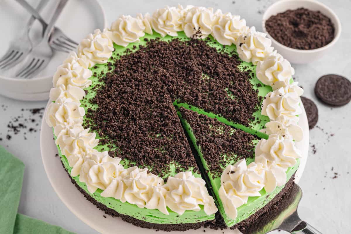 Grasshopper Cheesecake A round mint Grasshopper Cheesecake with a chocolate cookie crust, topped with green mint filling, whipped cream swirls around the edge, and crushed cookies in the center. One slice is being served. Plates, forks, and cookies are in the background.