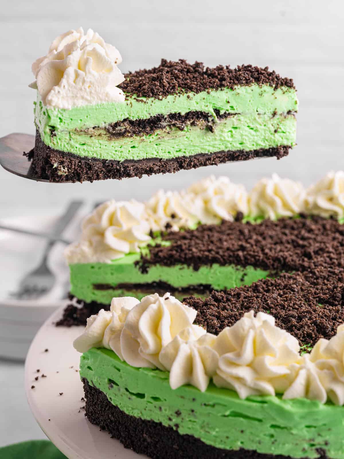 Grasshopper Cheesecake A slice of mint Grasshopper Cheesecake with a green creamy filling, chocolate cookie crust, chocolate crumbles on top, and piped whipped cream, being lifted from the whole pie on a white plate.