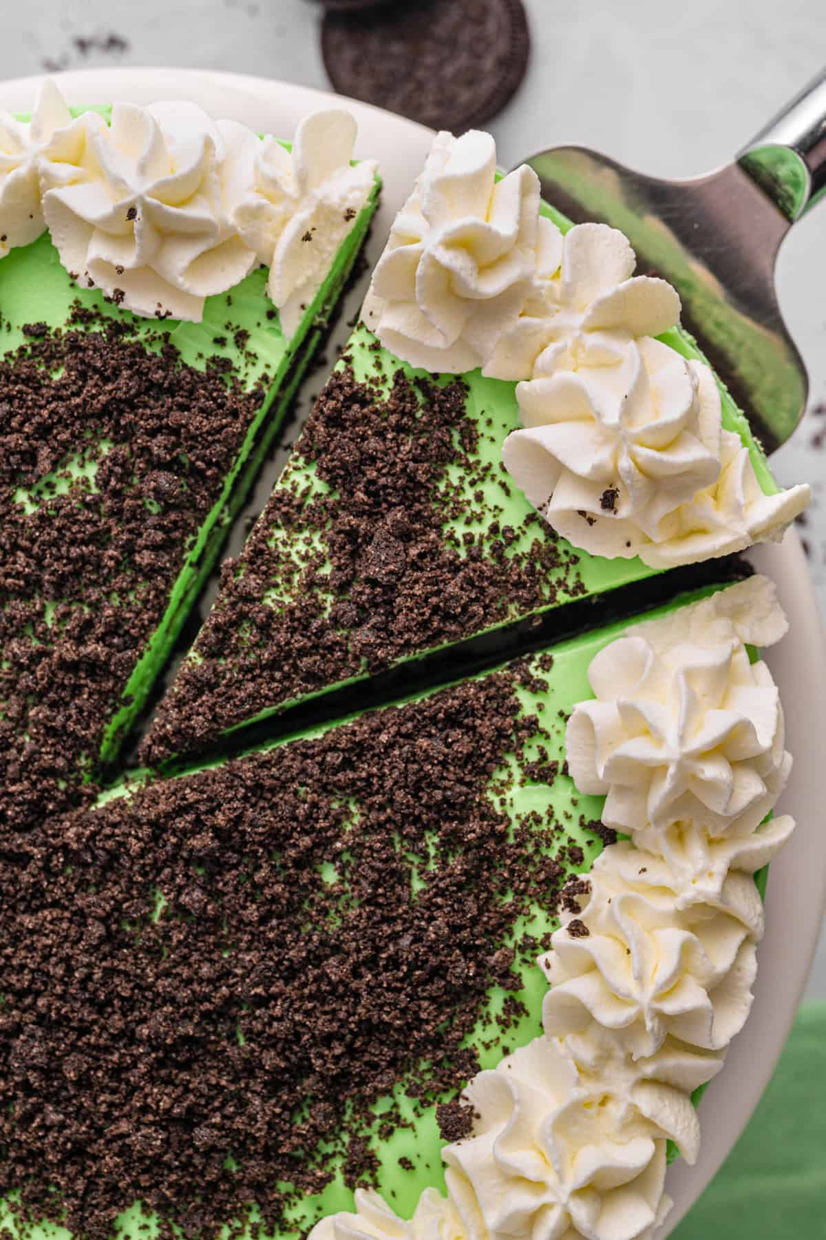 Grasshopper Cheesecake A top-down view of a green Grasshopper cheesecake with a slice being lifted. The cake is decorated with white whipped cream swirls around the edge and chocolate cookie crumbs scattered in the center.
