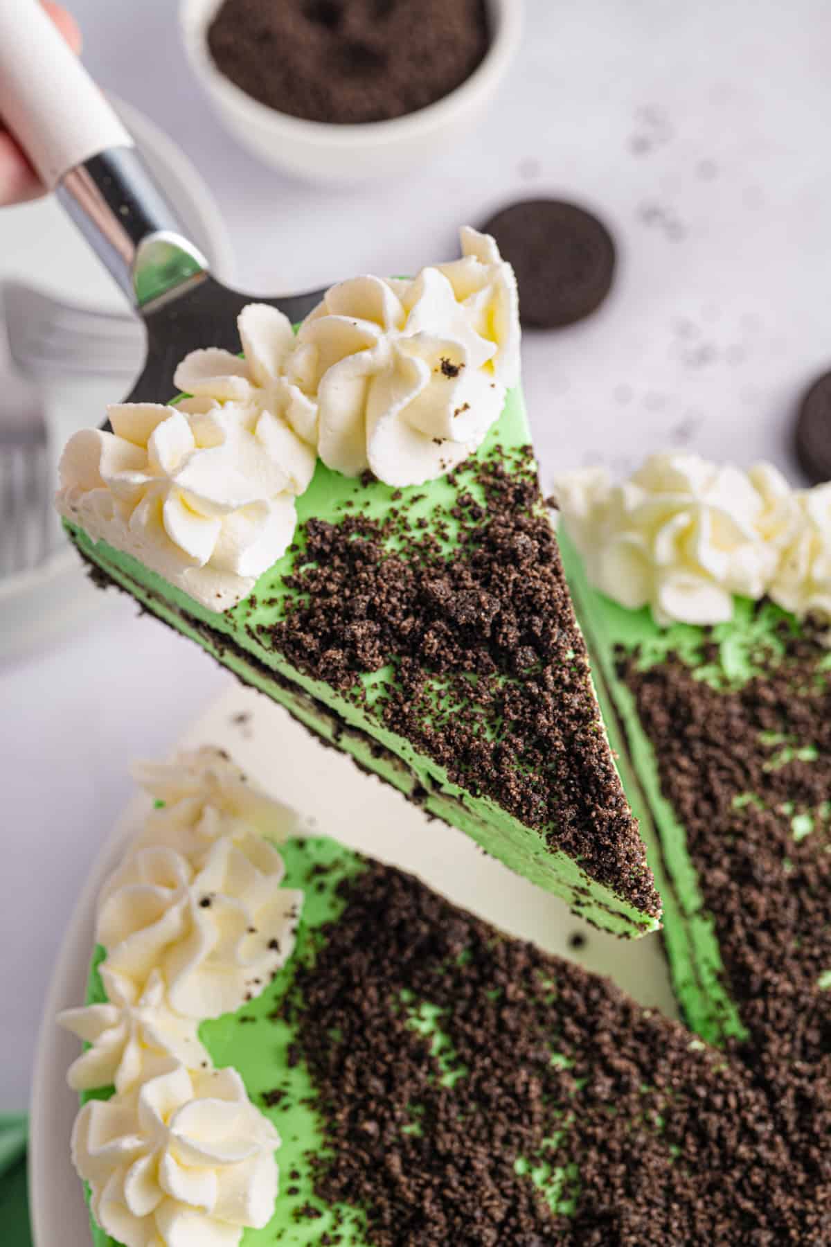Grasshopper Cheesecake A slice of green mint Grasshopper Cheesecake, topped with whipped cream rosettes and crushed chocolate cookies, is being lifted from the whole cake, which is decorated similarly. Bowls of cookies and crumbs are in the background.