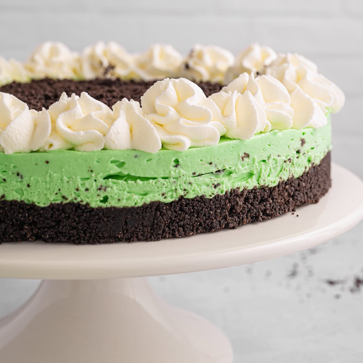 Grasshopper Cheesecake A Grasshopper Cheesecake with a green mint filling, topped with swirls of whipped cream, displayed on a white cake stand.