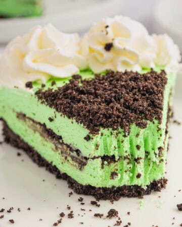 A slice of mint Grasshopper Cheesecake with whipped cream swirls on top, served on a white plate.