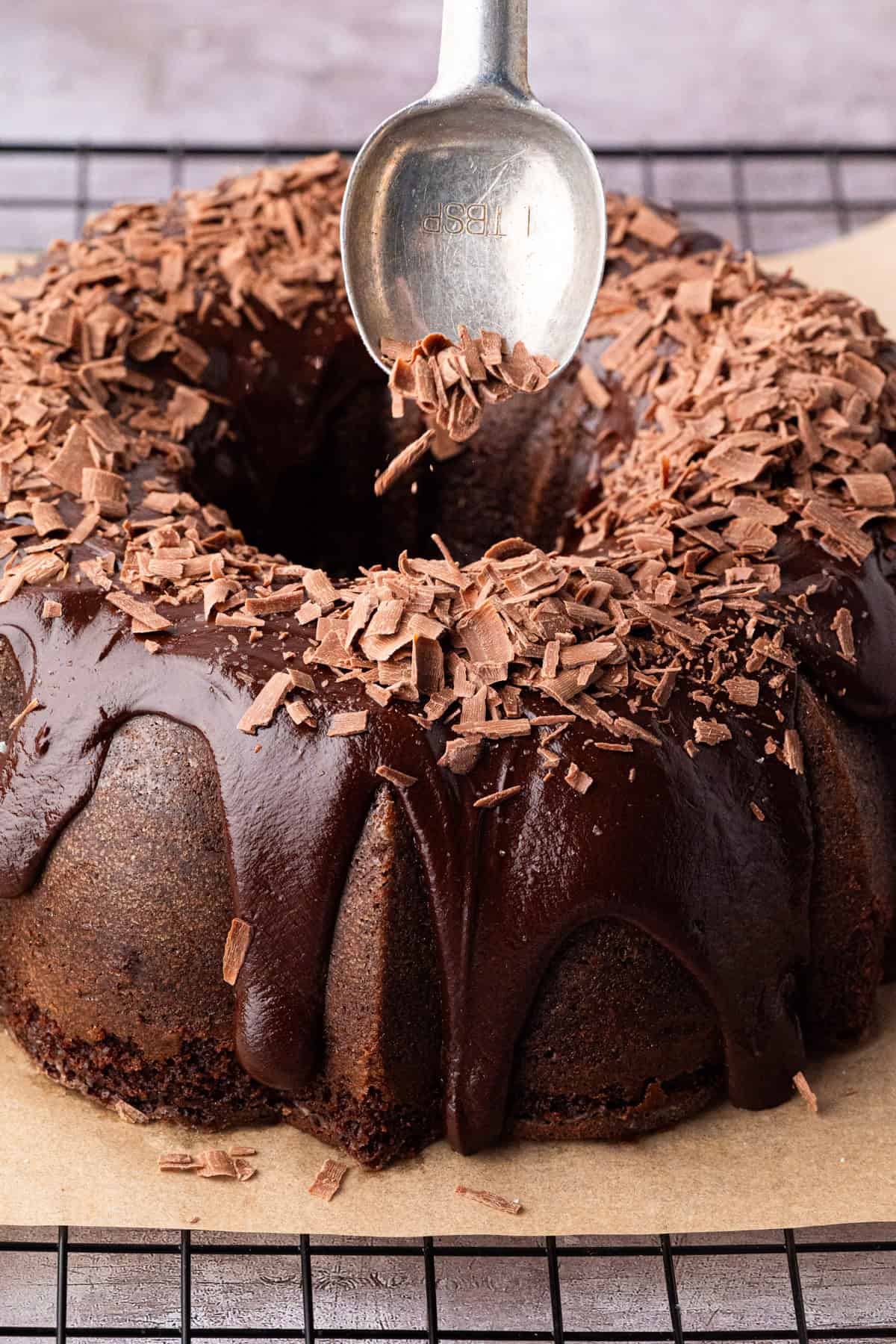 A chocolate bundt cake topped with glossy chocolate glaze and chocolate shavings, sits on parchment paper as a spoon sprinkles more shavings on top. 