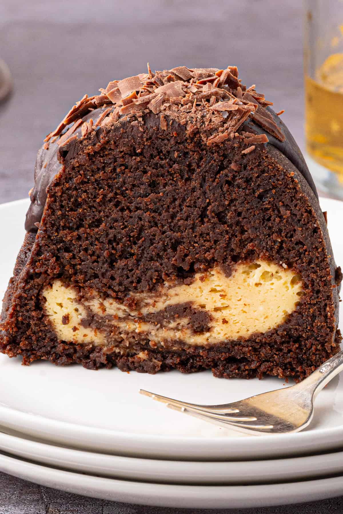 A slice of chocolate bundt cake with a creamy cheesecake filling, topped with chocolate shavings, sits on a stack of white plates next to a fork.