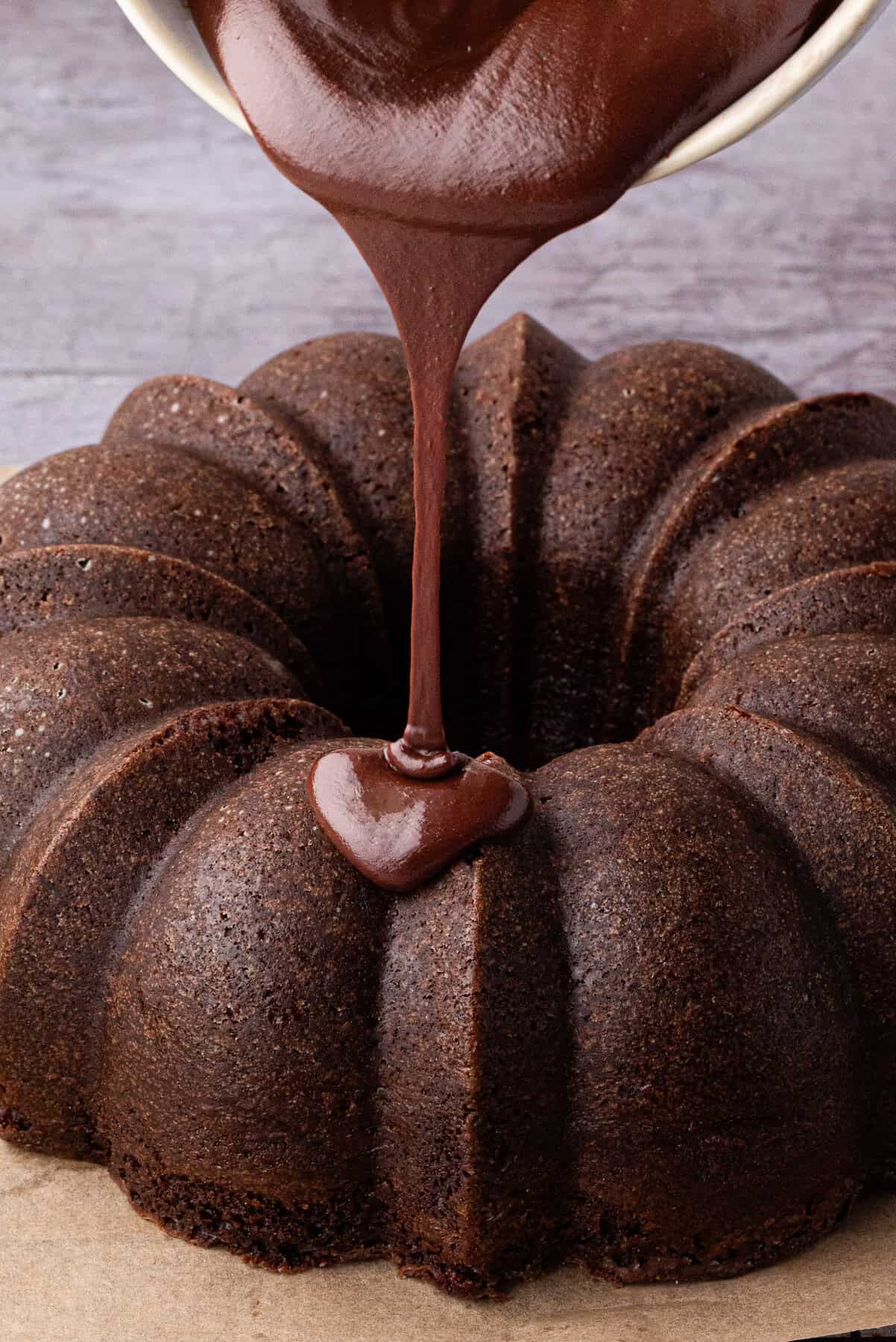 Chocolate glaze is being poured from a bowl onto the top of a round, ridged chocolate bundt cake. 