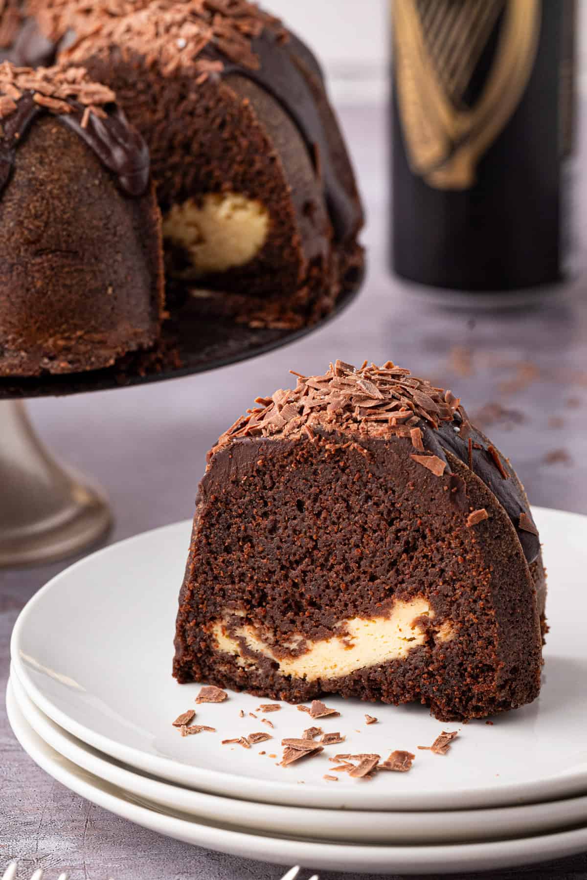 A slice of chocolate bundt cake with a creamy filling and chocolate shavings on top sits on a white plate, with the rest of the cake and a dark beverage can in the background.