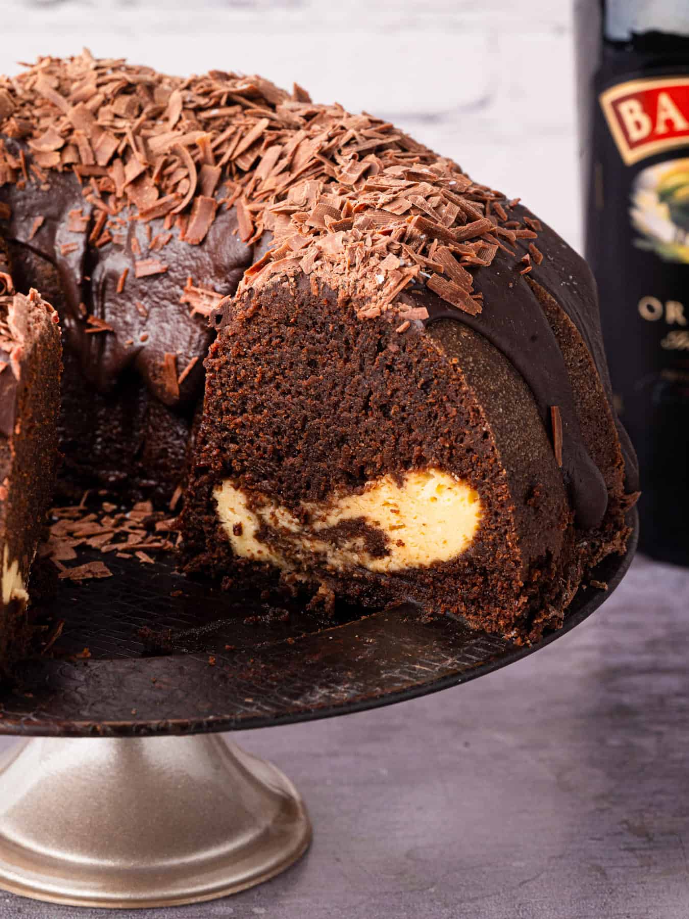 A chocolate Bundt cake with a cream cheese filling, topped with chocolate glaze and chocolate shavings, sits on a cake stand. A bottle of Baileys Irish Cream is visible in the background.