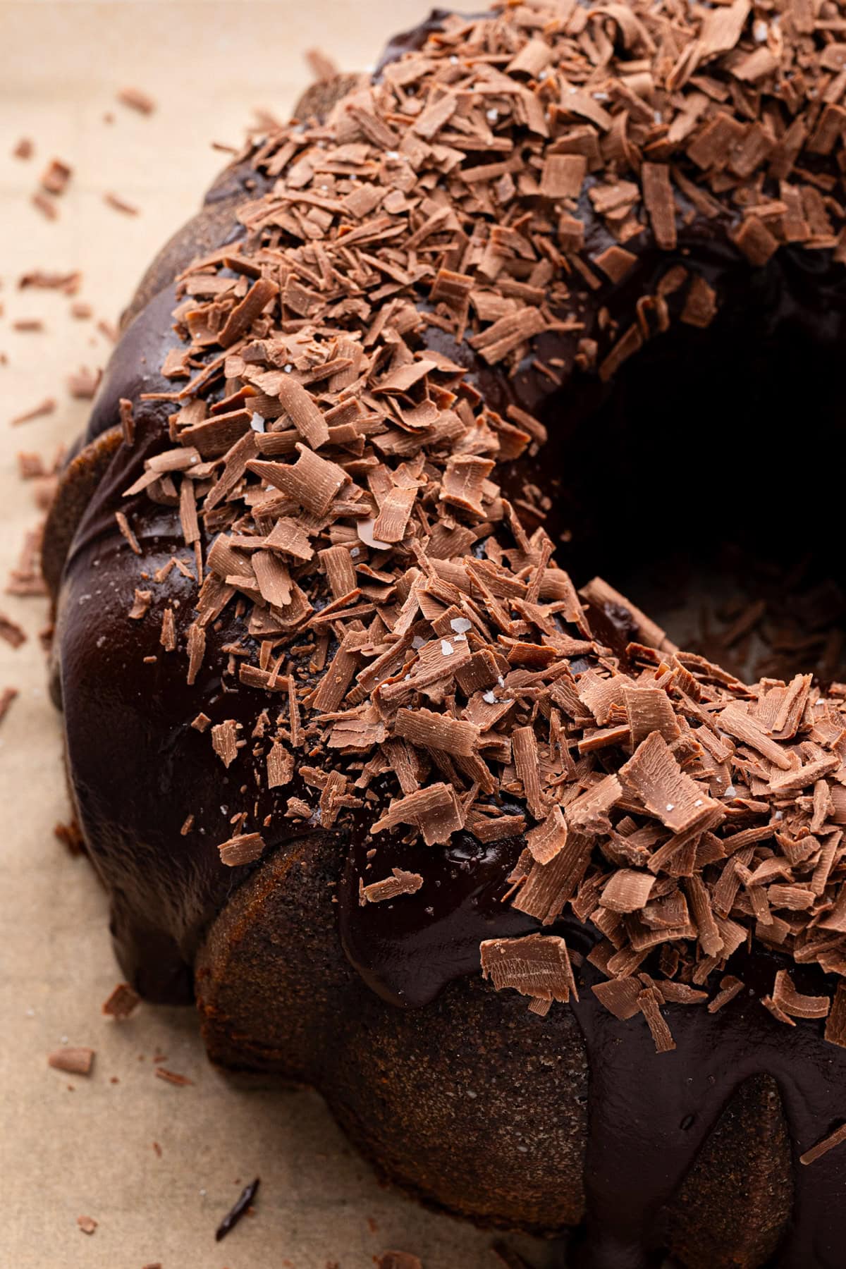 A close-up of a bundt cake topped with glossy chocolate glaze and generous chocolate shavings, set on a neutral background.