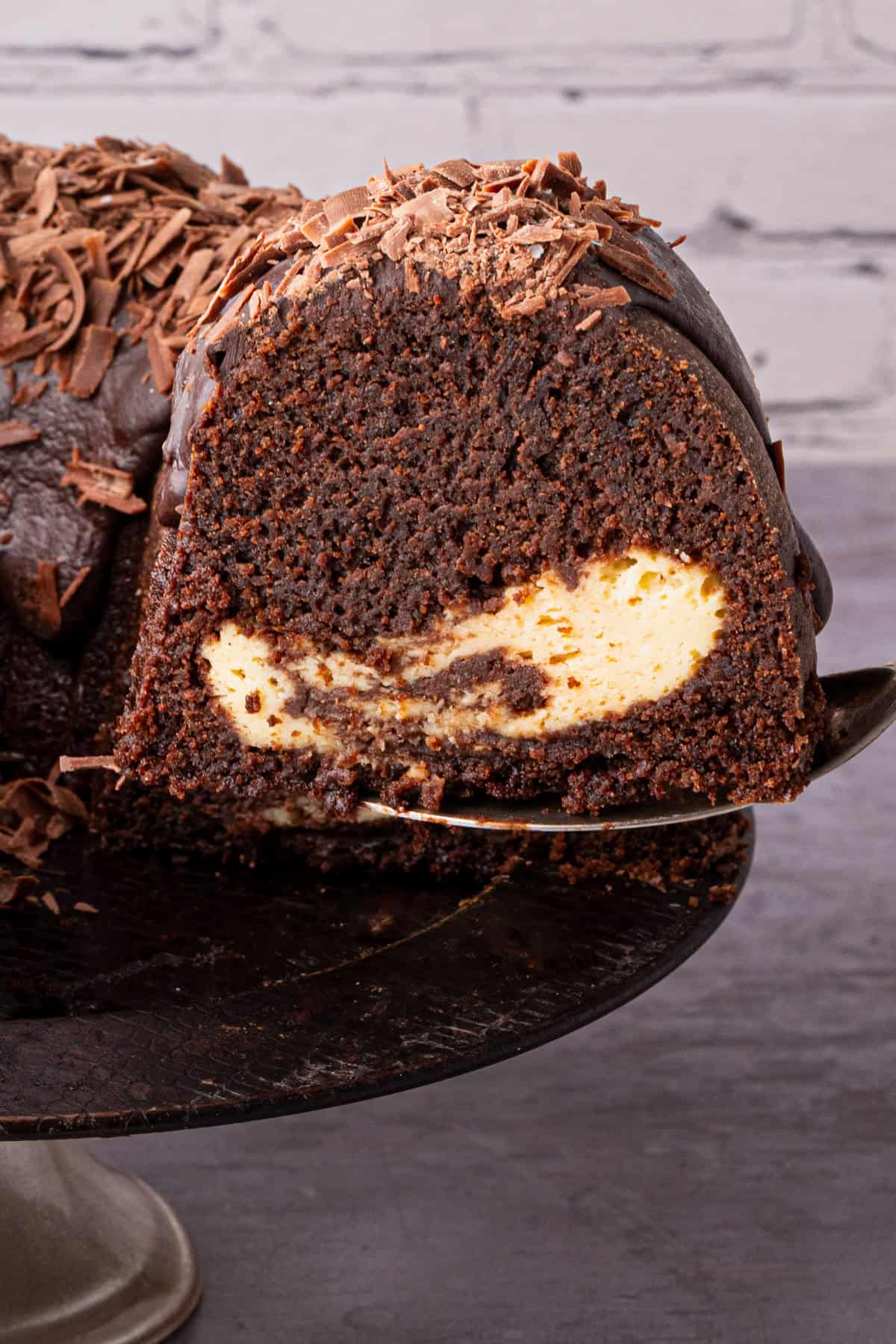 A close-up of a chocolate bundt cake with a creamy cheesecake filling, topped with chocolate shavings. A slice is being lifted from the cake, showing the rich, moist texture and contrasting filling inside.