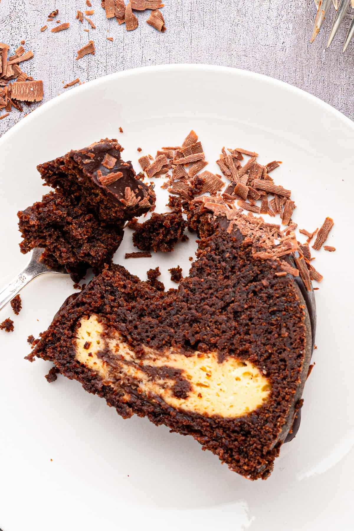 A slice of chocolate cake with a creamy filling and chocolate shavings sits on a white plate, with a fork beside it and cake crumbs scattered around.