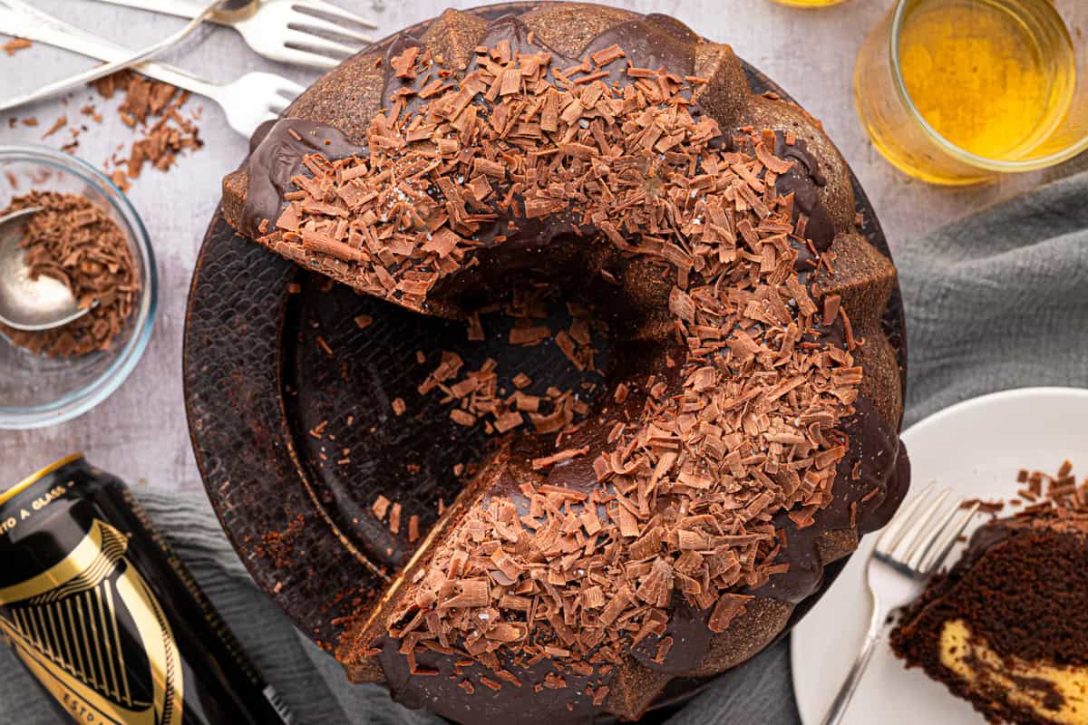 Overhead view of a chocolate bundt cake with chocolate shavings on top, partially sliced on a dark plate. Slices, forks, a can, and glasses are nearby on a gray surface.
