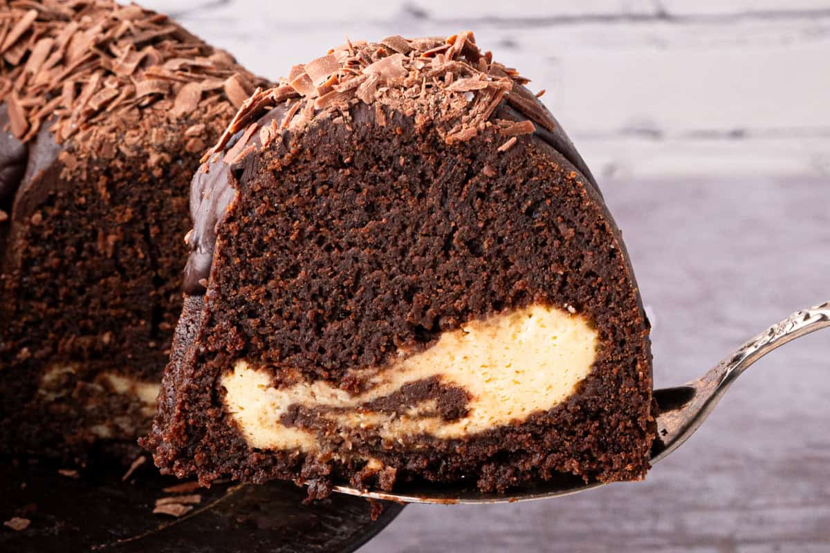 A close-up of a slice of chocolate bundt cake with a creamy cheesecake filling and chocolate shavings on top, held on a metal cake server.