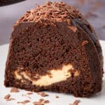 A slice of rich Guinness chocolate cake with a creamy Irish cream cheesecake filling in the center, topped with chocolate ganache and chocolate shavings, sitting on a white plate.