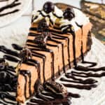 A slice of Mudslide Pie topped with whipped cream, chocolate drizzle, chocolate-covered espresso beans, and cookie pieces on a decorative white plate. Chocolate sauce is also drizzled on the plate.