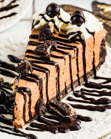 A slice of Mudslide Pie with Oreo crust, topped with whipped cream, chocolate drizzle, chocolate-covered espresso beans, and Oreo pieces, served on a white plate with extra chocolate sauce.