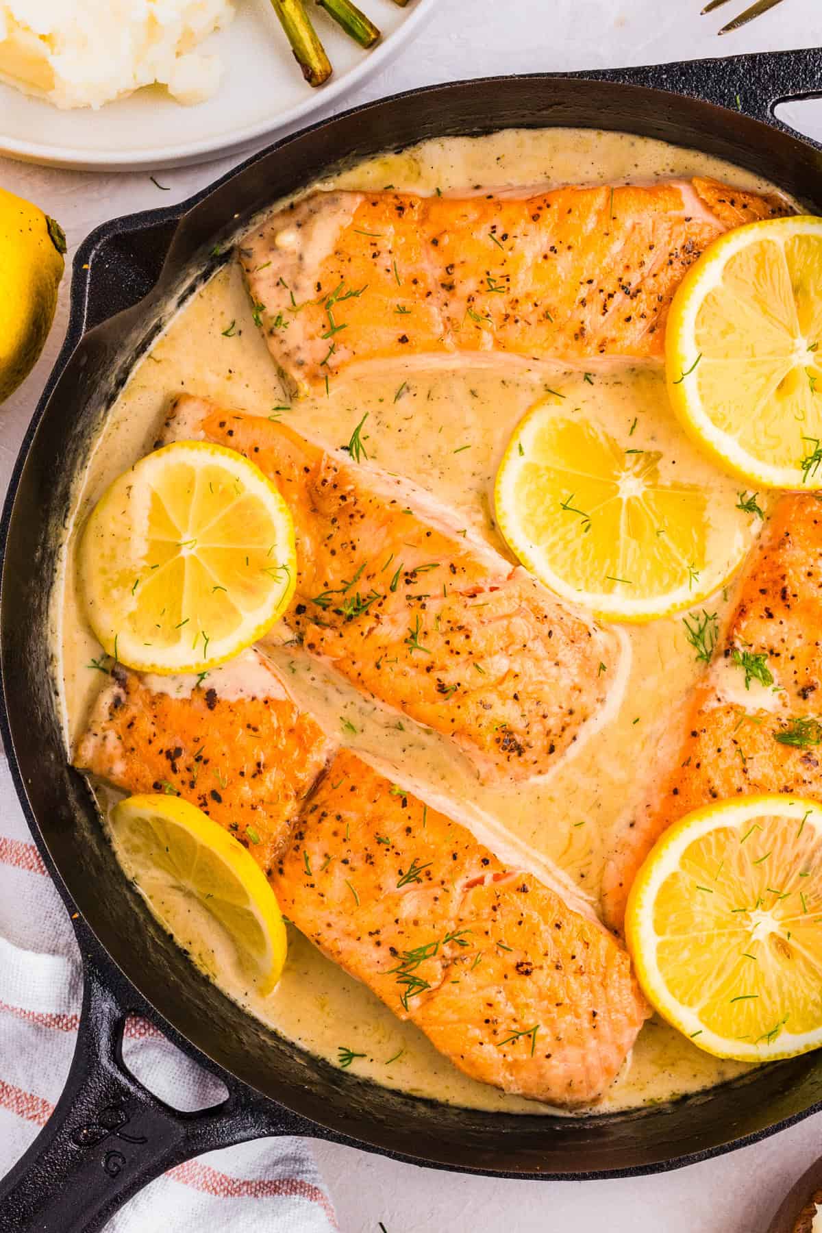 A skillet filled with cooked lemon dill salmon fillets in a creamy sauce, garnished with fresh dill and lemon slices. 