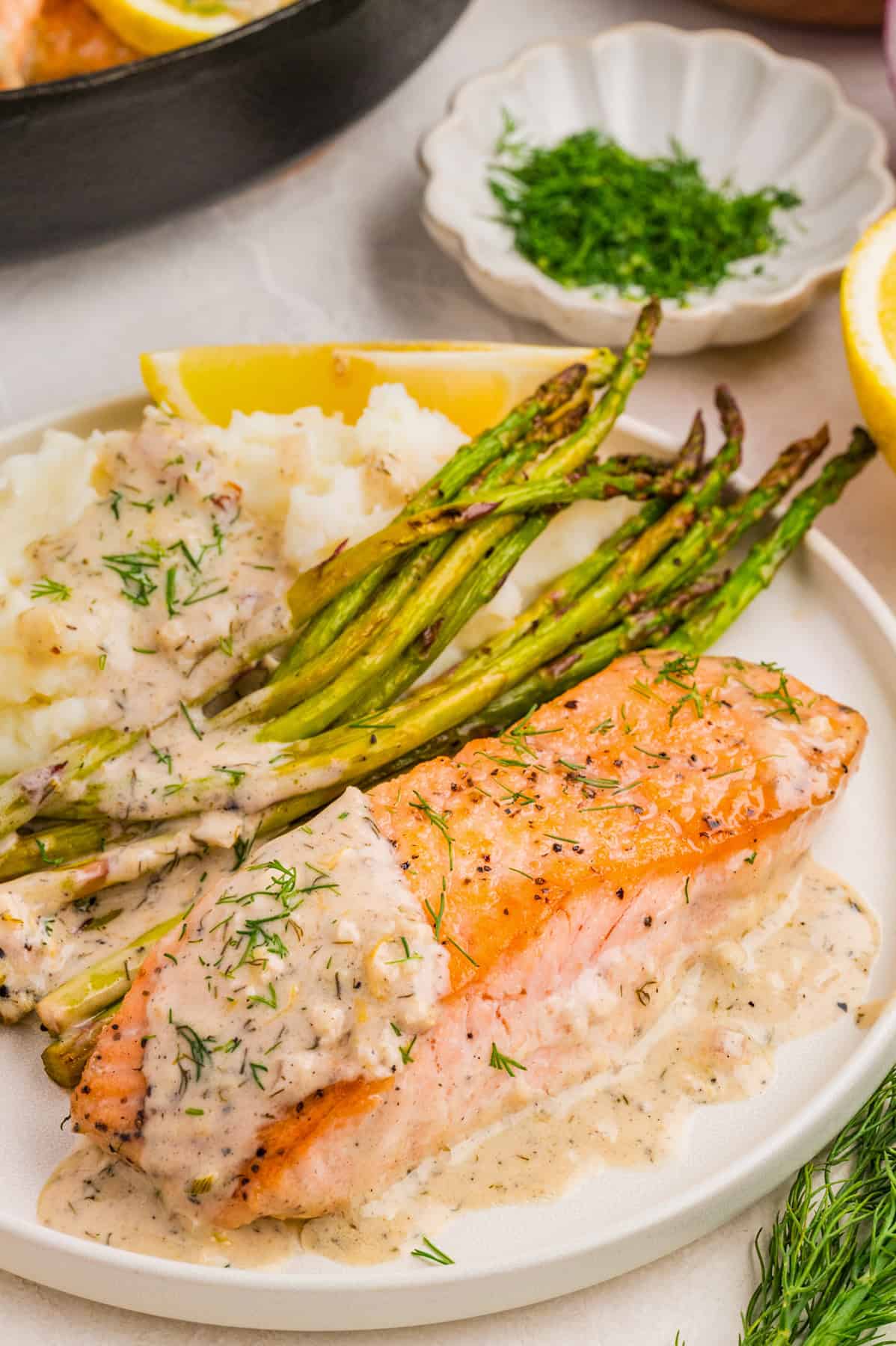 A plate with a cooked salmon fillet topped with creamy lemon dill sauce and fresh dill, served alongside mashed potatoes with roasted asparagus. 