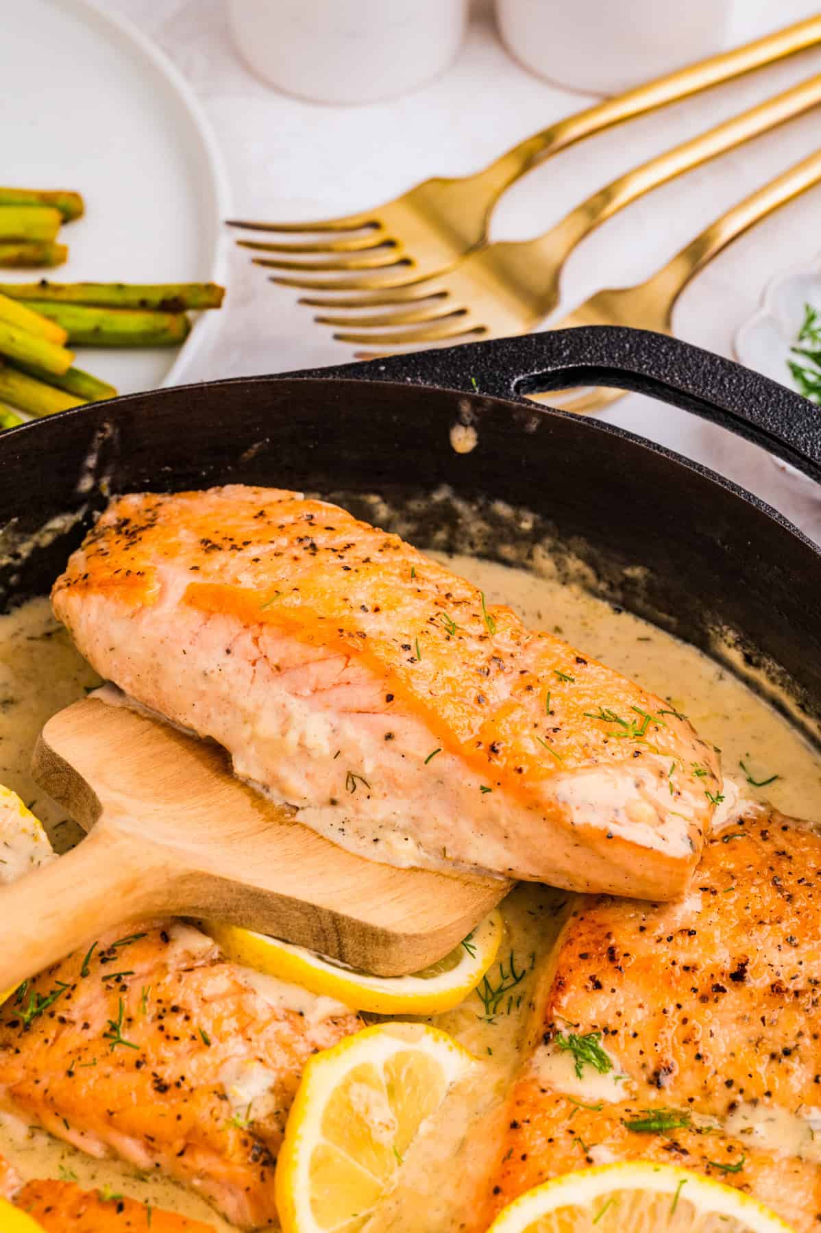 A skillet with creamy lemon dill sauce holds seared salmon fillets, garnished with dill and lemon slices. One fillet is lifted on a wooden spatula.