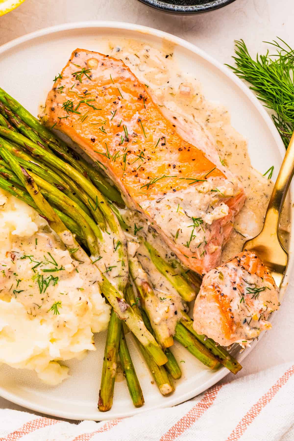 A plate with lemon dill salmon in creamy sauce, mashed potatoes, and cooked asparagus, garnished with fresh dill. A fork holds a bite of salmon.