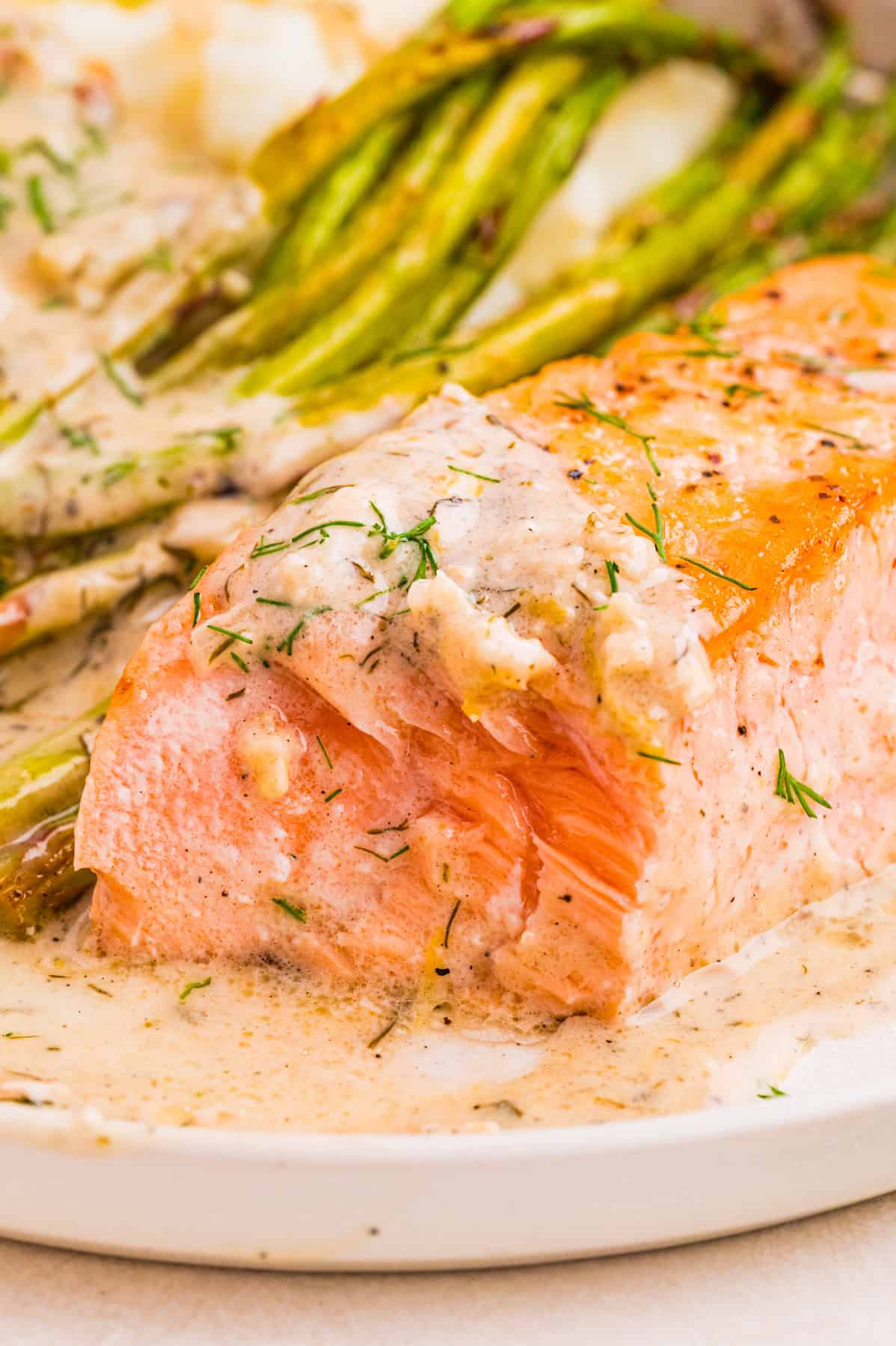 A piece of cooked salmon topped with creamy lemon dill sauce sits on a plate next to roasted asparagus and mashed potatoes.