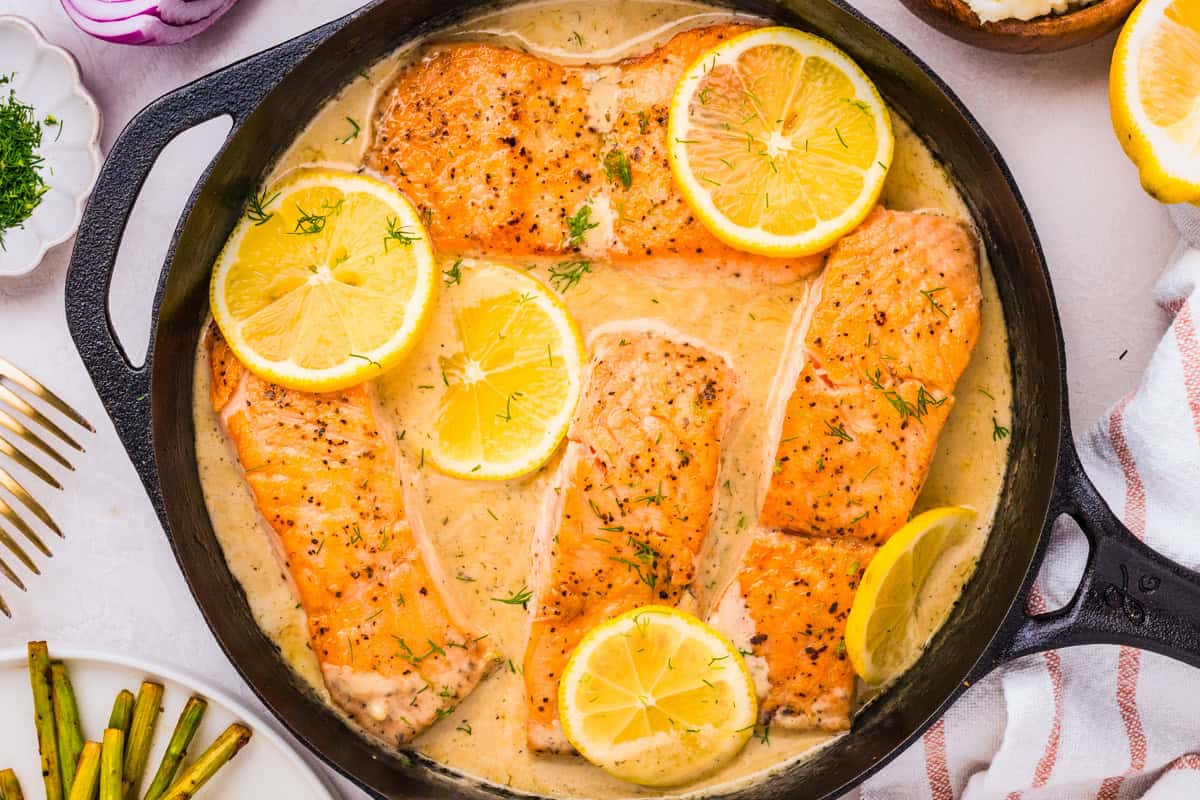 Four salmon fillets in a creamy lemon dill sauce are cooked in a black skillet, topped with lemon slices and sprinkled with herbs. Nearby, there are lemon halves, asparagus, and a striped towel.