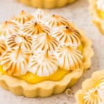 A close-up of a lemon meringue tartlet topped with toasted meringue peaks, set on a light-colored surface.