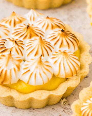 A close-up of a lemon meringue tartlet topped with toasted meringue peaks, set on a light-colored surface.