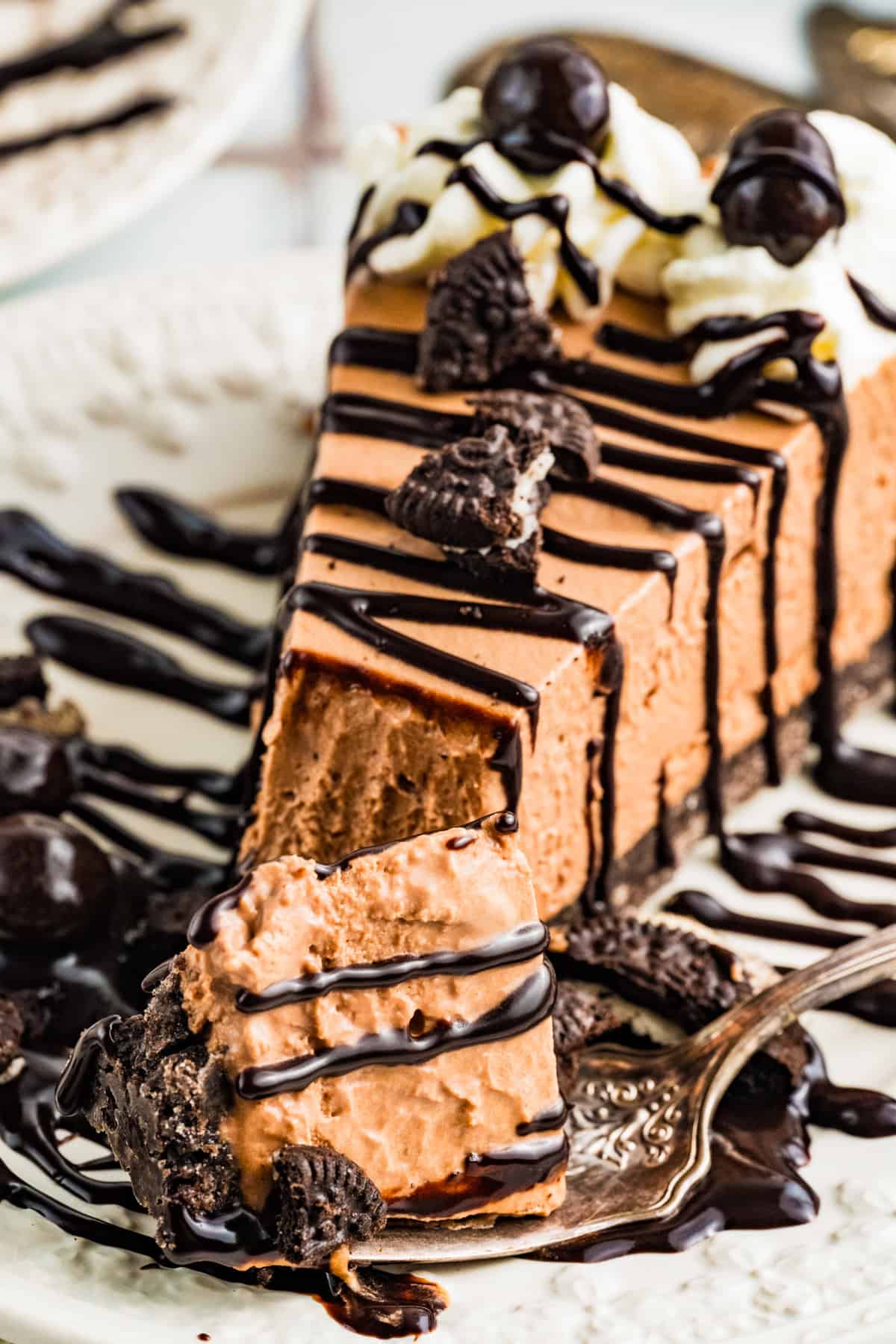 Mudslide Pie A slice of Mudslide Pie topped with whipped cream, chocolate cookies, and chocolate espresso beans, drizzled with chocolate sauce. A fork holds a bite of the creamy cheesecake in the foreground.