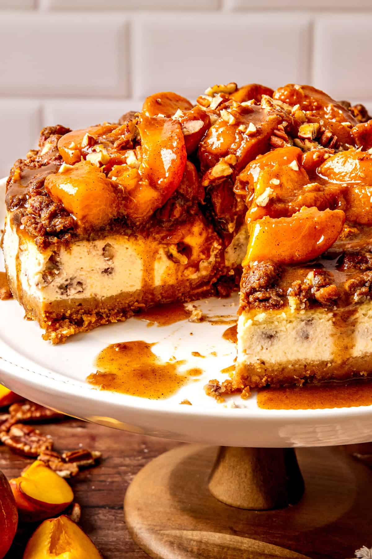 A peach cobbler cheesecake topped with glazed peaches and chopped nuts sits on a white cake stand. A large slice has been removed, revealing a creamy interior with visible chunks and a crumbly brown crust.