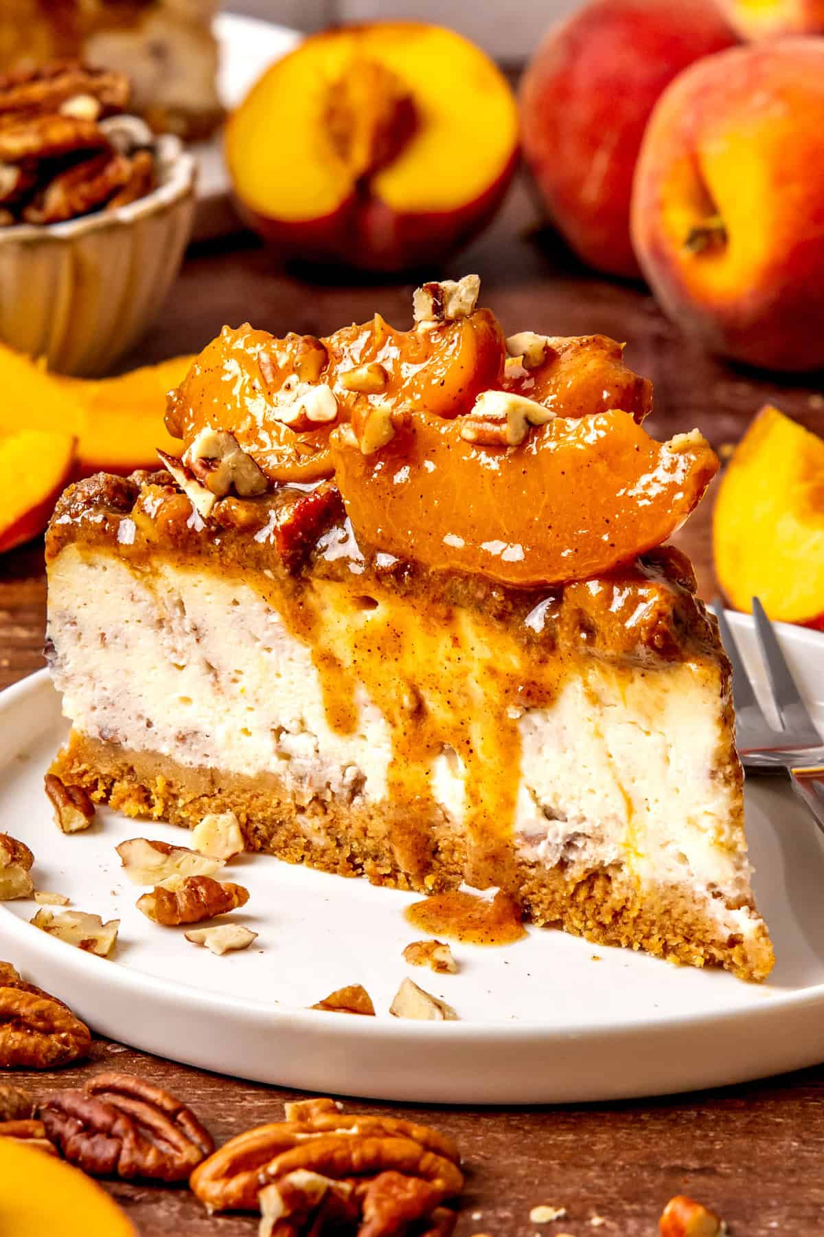 A slice of peach cobbler cheesecake topped with caramelized peach slices and chopped pecans on a white plate, surrounded by fresh peaches and pecans on a wooden table.