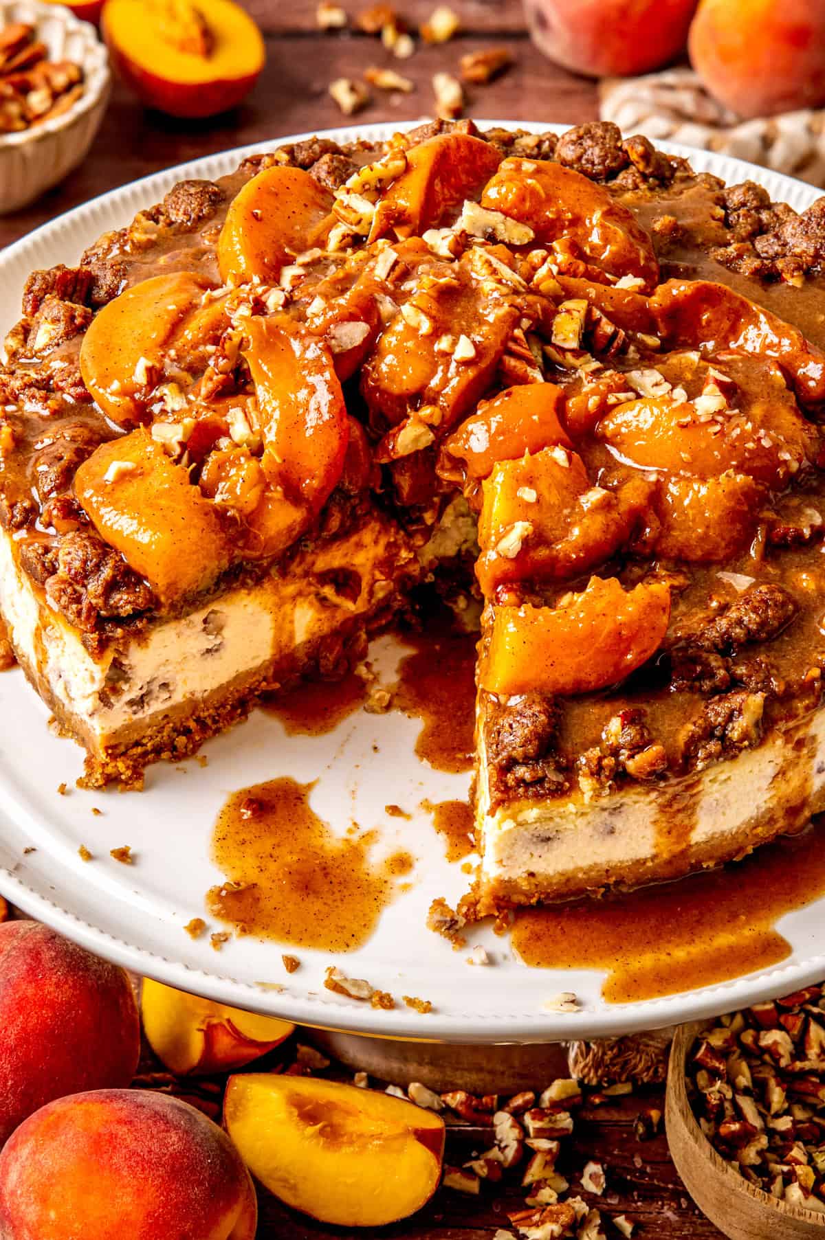 A Peach Cobbler Cheesecake topped with caramelized peach slices and chopped pecans sits on a white plate, one slice removed. Fresh peaches and pecans are scattered around on the wooden table for a rustic touch.