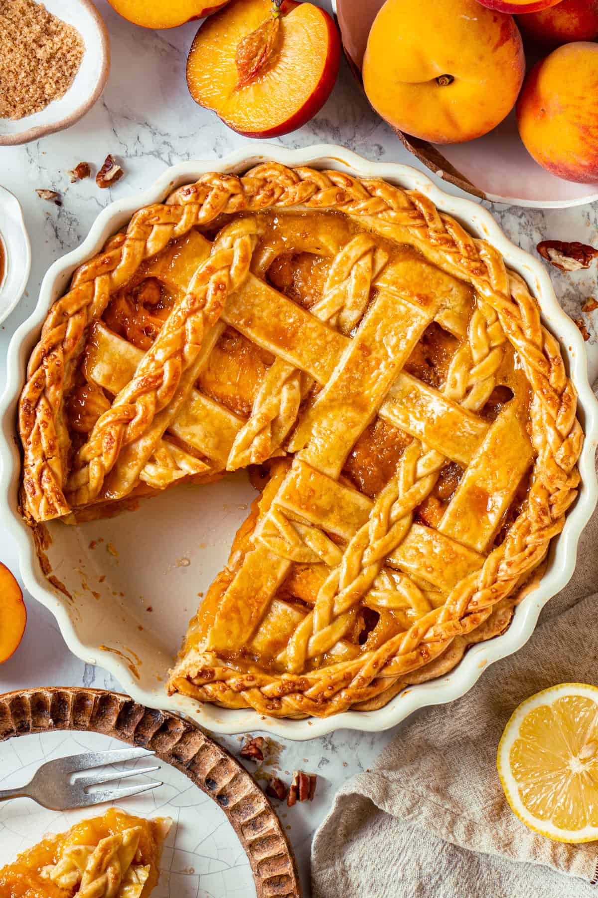 A Peach Pie with a golden lattice and braided crust sits in a white dish, with a slice missing. Fresh peaches, a lemon slice, and a plate holding a slice of peach pie are arranged on the marble surface nearby.
