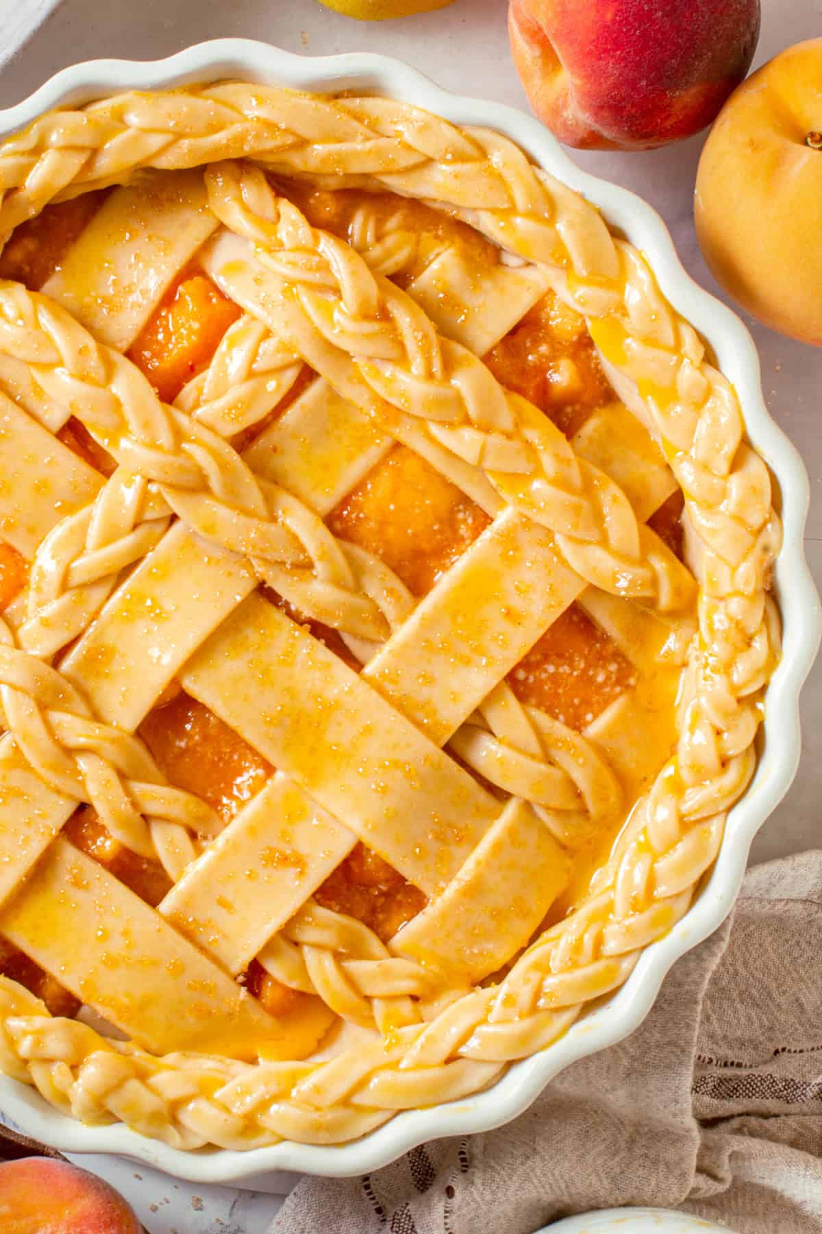 An unbaked Peach Pie with a lattice and braided crust sits in a white pie dish, surrounded by whole peaches on a light background.