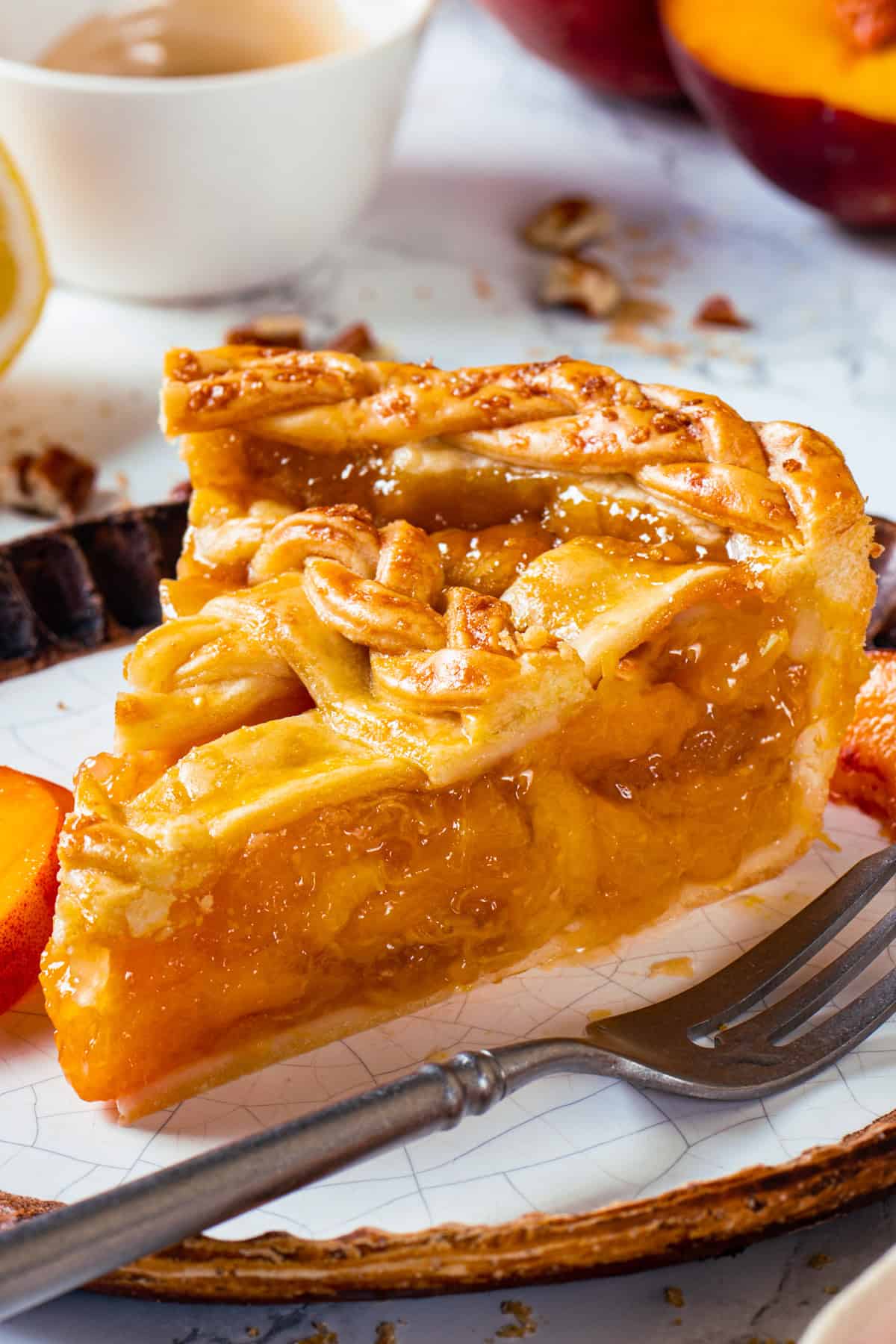A close-up of a slice of Peach Pie with a golden, flaky crust and glossy peach filling on a plate, next to a fork. A cup and fresh peach slices are visible in the background.