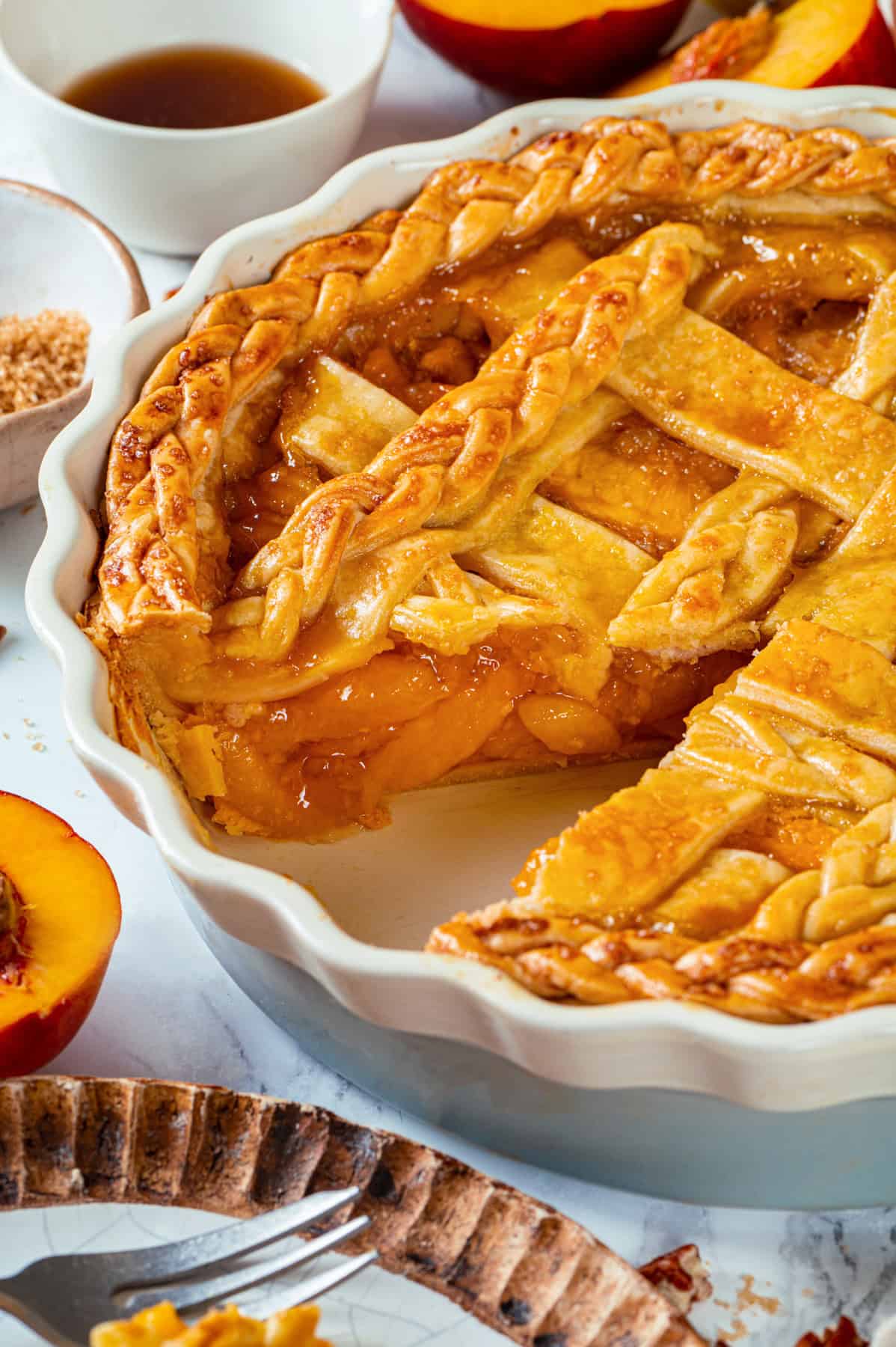 A close-up of a Peach Pie with a golden, braided lattice crust in a white dish. A slice is missing, revealing juicy peach filling. Surrounding the pie are fresh peach halves, a cup of sauce, and a dish of brown sugar.