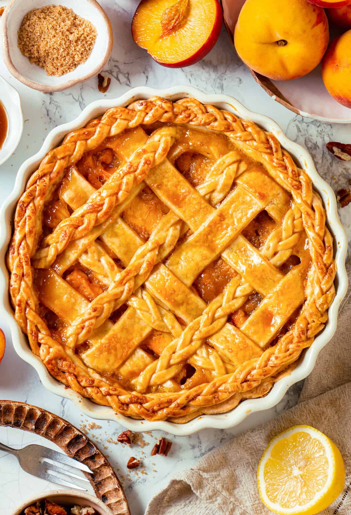 A golden-brown Peach Pie with a lattice and braided crust sits in a white dish, surrounded by fresh peaches, a lemon half, brown sugar, and pie slices on a marble surface.