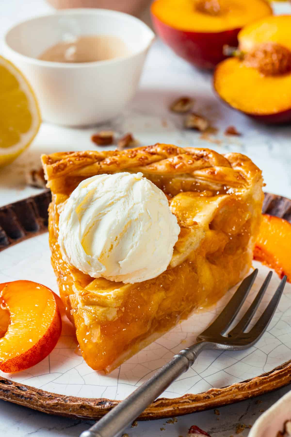 A slice of peach pie topped with vanilla ice cream sits on a plate with fresh peach slices, a fork, and a blurred background featuring more peaches and a lemon half.