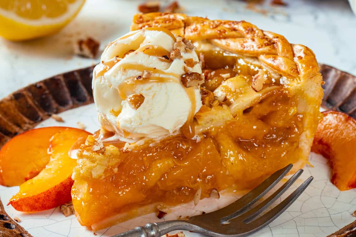 A slice of peach pie topped with vanilla ice cream, caramel drizzle, and chopped nuts is served on a plate with fresh peach slices and a fork.