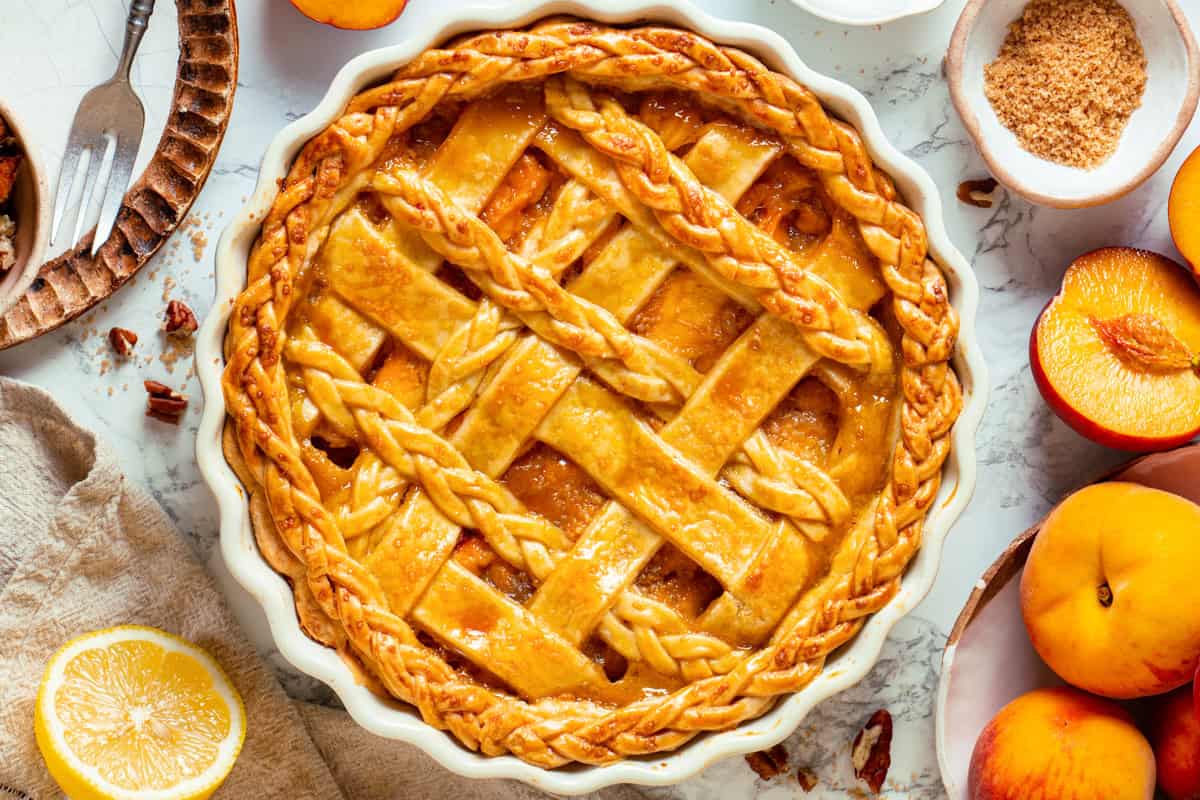 A golden-brown Peach Pie with a decorative lattice crust sits in a white pie dish, surrounded by fresh peaches, a halved lemon, a bowl of brown sugar, and a fork on a marble surface.