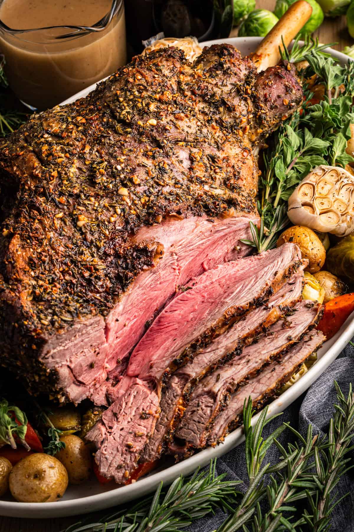 A large, herb-crusted roast leg of lamb is sliced to reveal a juicy pink interior. The platter is garnished with fresh herbs, roasted potatoes, garlic, and carrots, with a bowl of brown gravy in the background.