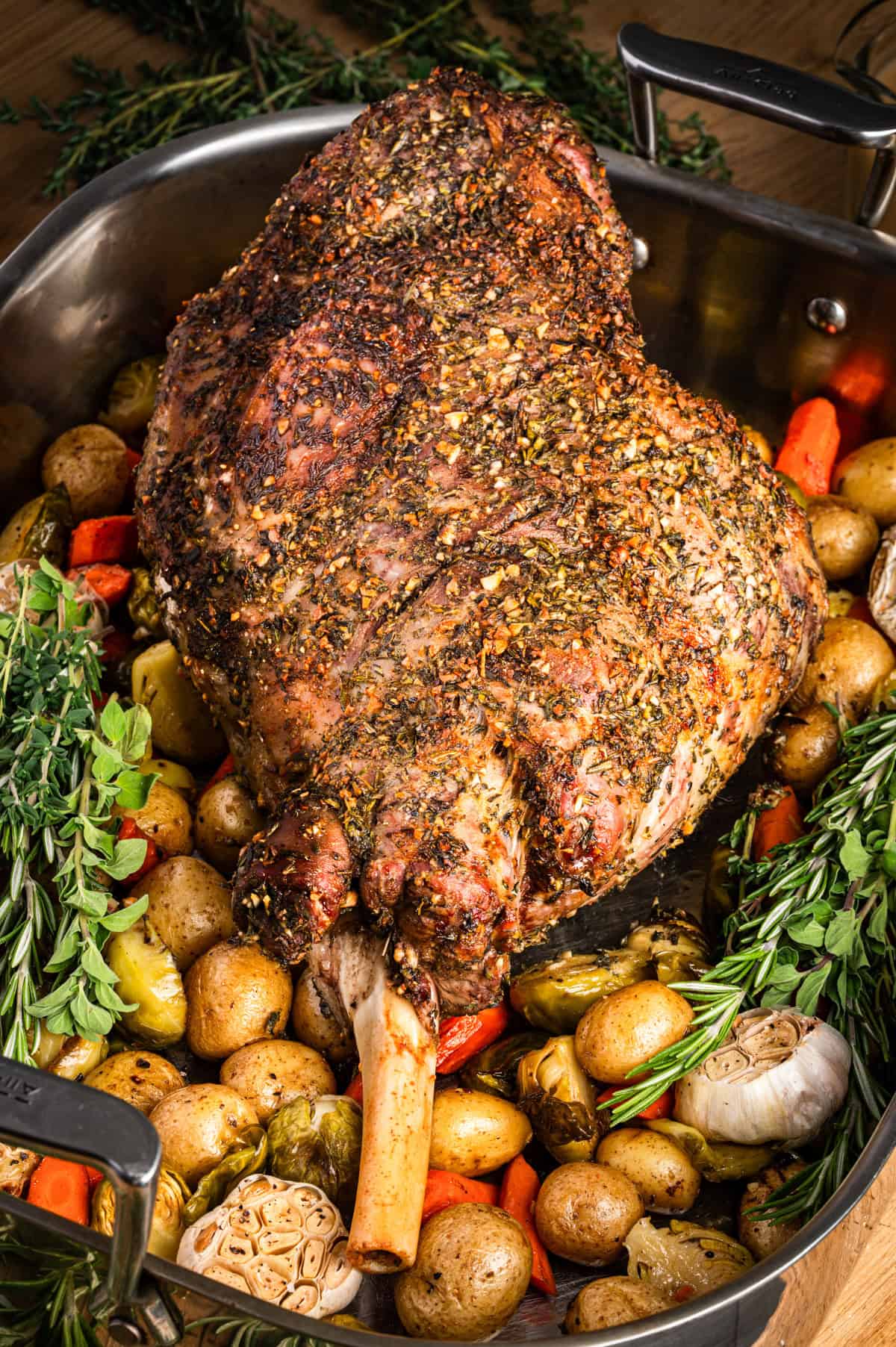 A seasoned, roasted leg of lamb sits in a roasting pan surrounded by baby potatoes, carrots, garlic, brussels sprouts, and fresh herbs. 